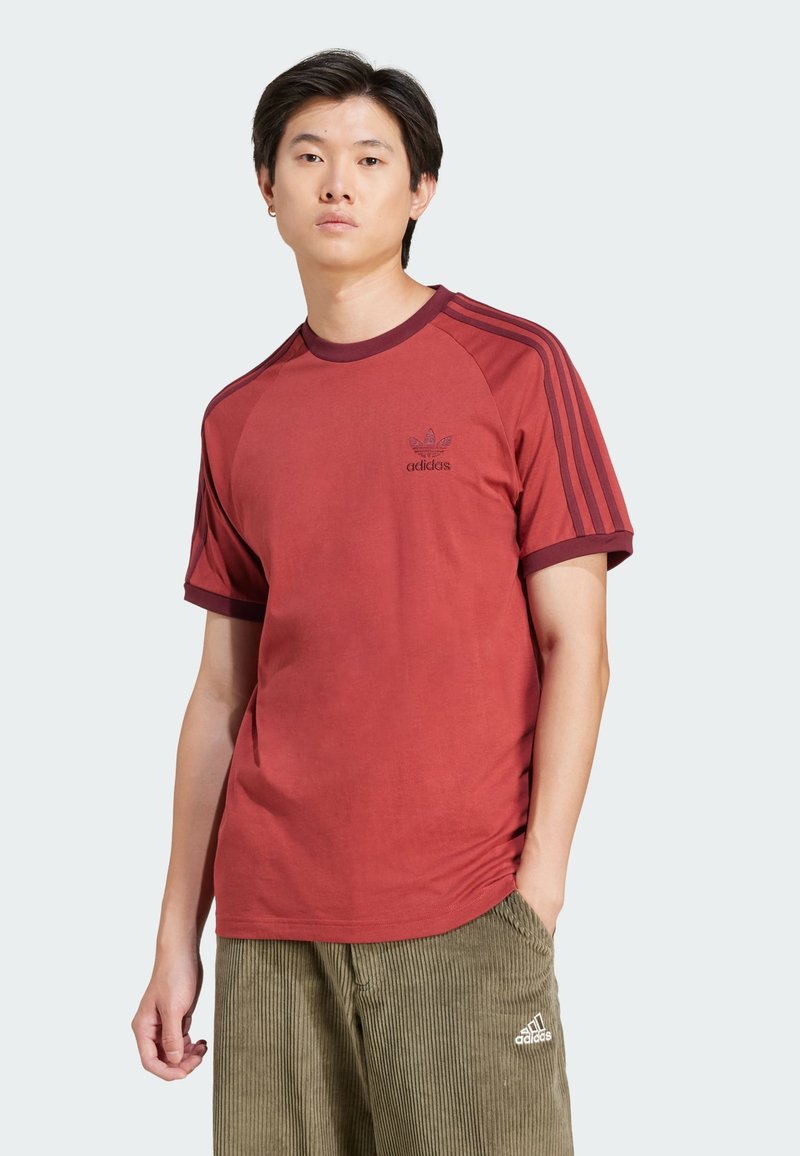 Crvena atletska majica kratkih rukava, rebrastog ovratnika, s tri crne pruge na ramenima. Na prsima se nalazi mali crni adidas logotip.
