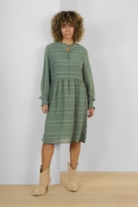 Grön långärmad klänning med knäppning och volangkrage; har randigt mönster; matchad med beige ankelboots på ett trägolv.
