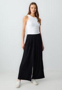 Pantalon noir à jambes larges avec plis, associé à un débardeur blanc ajusté. La tenue présente une texture lisse et des baskets blanches décontractées.