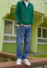 Zelená mikina Nike na zip, pod ní bílý tričko, modré džínové kalhoty a bílé tenisky Nike. Na pozadí zelená budova.