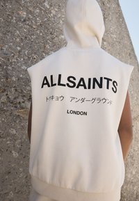 Felpa senza maniche color crema con un grande logo nero "ALLSAINTS" sul retro, con testo giapponese e "LONDON" sotto, realizzata in morbido cotone.