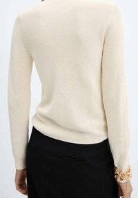 Pull en maille côtelée ivoire à manches longues. Le dos est sans couture. Associé à une jupe noire taille haute accentuée par un bracelet en chaîne dorée.