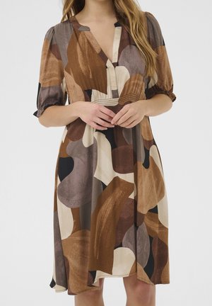 Femme portant une robe jusqu'aux genoux avec des formes abstraites marron, beige et noires, manches bouffantes arrivant au niveau des coudes et décolleté en V, mains jointes à la taille.