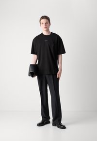 Černé oversized tričko s decentním logem a zipovou kapsou, v kombinaci s černými tailorovanými kalhotami a lesklými černými botami.