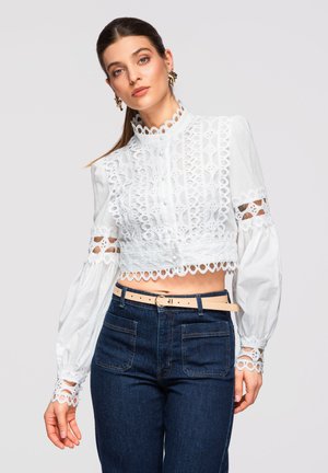 Kobieta ubrana w białą koronkową, krótką bluzkę z bufkami, niebieskie jeansy z wysokim stanem i jasnobeżowy pasek na szarym tle.