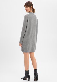 Szara dzianinowa sukienka swetrowa z wysokim kołnierzem, długimi rękawami, rozcięciami po bokach i prążkowanym dołem. Noszona z czarnymi botkami za kostkę na jednolitym tle.
