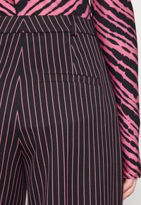 Černé kalhoty s vertikálními růžovými pruhy, obsahující boční kapsu a hladkou texturu. Nosené s dlouhým rukávem ve kontrastním vzoru.