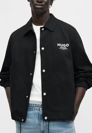 Sort knap-op jakke med hvidt "HUGO" logo på brystet, båret over hvid skjorte og lyseblå jeans, hænder delvist i lommerne.