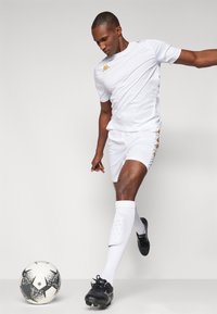 Kit atlético blanco con detalles en dorado, que incluye una camiseta de manga corta y pantalones cortos. Zapatillas de fútbol negras y calcetas blancas hasta la rodilla con textura.