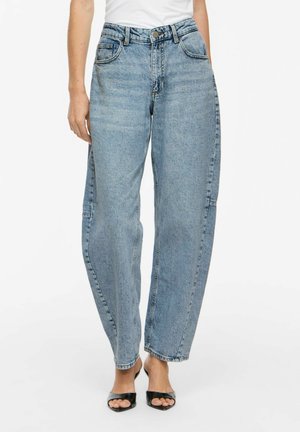 Person iført lyseblå jeans med høj talje og løs pasform samt sorte åbne hælede sandaler, stående foran en hvid baggrund.
