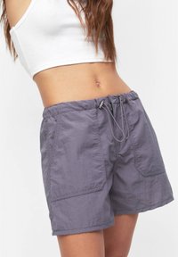 Nainen, jolla on harmaat nyöritettävät shortsit ja valkoinen lyhyt toppi, näkyy puolivälistä vartaloa keski-reiteen asti yksinkertaista taustaa vasten.