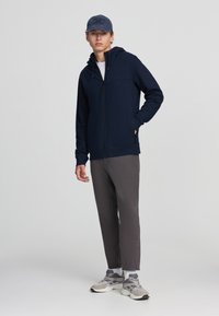 Felpa con zip di colore navy con finitura testurizzata, dotata di cappuccio e due tasche laterali. Abbinata a pantaloni grigi e scarpe sportive grigie chiare.