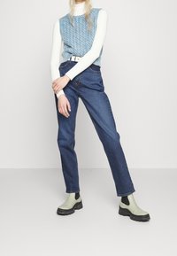 Top de punto sin mangas en azul claro texturizado, camiseta de manga larga blanca con canalé, jeans azul oscuro y botas de tobillo verdes con acentos negros.