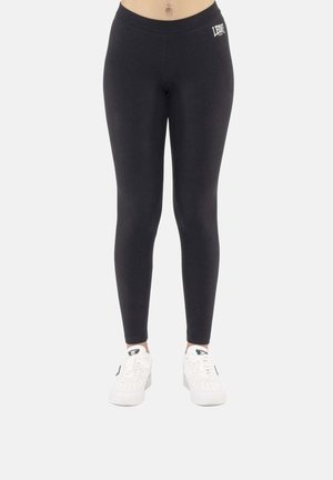 Leggings in Schwarz mit Logo an der linken Hüfte, getragen von einer Person, kombiniert mit weißen Sneakers, vor hellem einfarbigem Hintergrund.