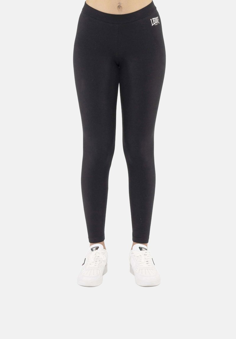 Leggings noirs avec logo sur la hanche gauche portés par une personne, associés à des baskets blanches, sur un fond clair uni.