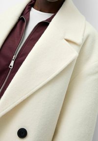 Roomkleurige wollen jas met een gestructureerde kraag en zwarte knoop, gecombineerd met een bordeaux rode ritsjas, waaronder een wit T-shirt met ronde hals zichtbaar is.