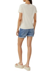 Crème katoenen t-shirt en blauwe denim shorts met opgerolde mouwen. Persoon draagt beige platte sandalen. Achteraanzicht, minimalistisch ontwerp.