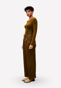 Jonge vrouw met kort krullend haar, gekleed in een aansluitend bruin langemouwen shirt, hoge taille wijde bruine broek en beige loafers, staande in zijaanzicht.