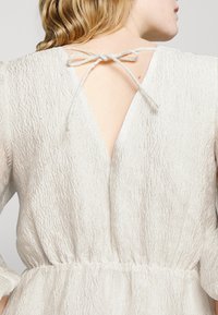 Γυναίκα με ξανθά μαλλιά που φοράει ένα υφασμάτινο λευκό φόρεμα με V-κοπή στην πλάτη, δεμένο με φιόγκο στον λαιμό.