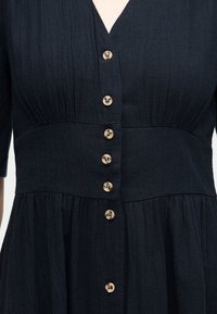 Σκούρο ναυτικό φόρεμα με κοντά μανίκια, V-λαιμόκοψη, σούρα στη μέση, υφασμάτινη υφή και μια κατακόρυφη σειρά από οκτώ ανοιχτό καφέ κουμπιά μπροστά.