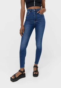 Jeans skinny de denim azul de talle alto con diseño de cinco bolsillos, detalles de costura y una textura suave. Llevados con sandalias de plataforma negras.