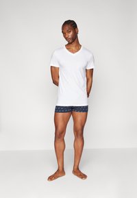 T-shirt blanc à col en V associé à un short en boxeur bleu à motifs. Le short présente un petit motif répété et un ajustement serré. Le modèle est pieds nus.