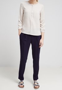 Blusa beige chiaro con maniche lunghe e dettagli con bottoni, abbinata a pantaloni plissettati navy scuro e sandali con cinturini metallici.