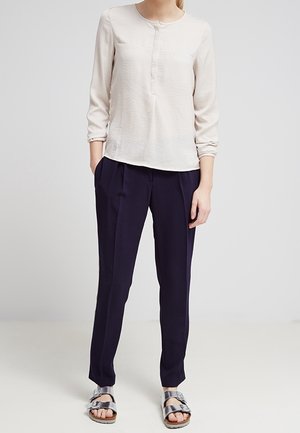 Blusa beige chiaro con maniche lunghe e dettagli con bottoni, abbinata a pantaloni plissettati navy scuro e sandali con cinturini metallici.