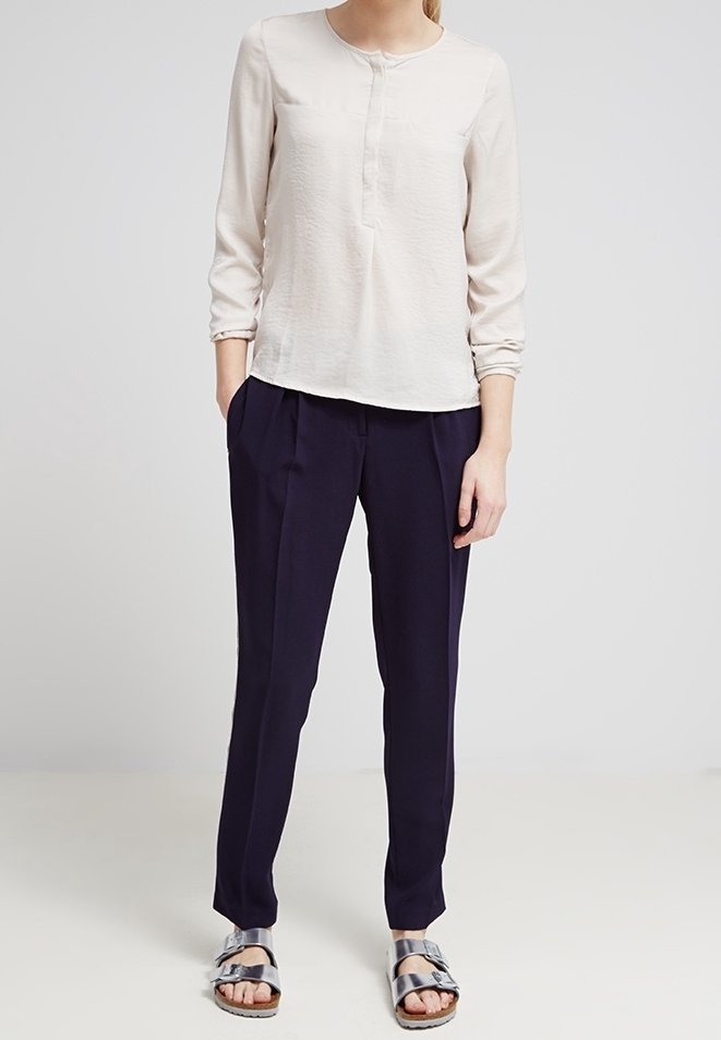 Blusa beige chiaro con maniche lunghe e dettagli con bottoni, abbinata a pantaloni plissettati navy scuro e sandali con cinturini metallici.