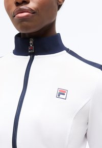 Άτομο που φοράει λευκό μπουφάν με φερμουάρ, με ναυτικό γιακά και ώμους, με κόκκινο και ναυτικό λογότυπο στο αριστερό μέρος του στήθους.