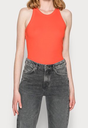 Vrouw met een mouwloos feloranje aansluitend topje en hooggetailleerde vervaagde zwarte jeans tegen een effen achtergrond.