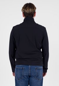 Giacca softshell nera con colletto alto, design snello, maniche lunghe e dettagli di cucitura discreti, abbinata a jeans in denim blu.