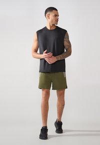 Juodas be rankovių marškinėliai derinami su alyvuogių spalvos šortais. Apranga pasižymi lygiu paviršiumi ir juodais sportiniais batais su minimaliais akcentais.