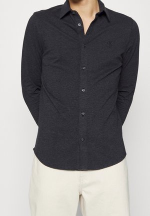 Homme portant une chemise charbon foncé à manches longues avec boutons et col, et un pantalon beige clair sur un fond uni.