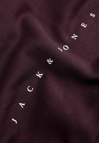 Material burgundy cu o textură netedă, având numele brandului "JACK & JONES" în alb, poziționat vertical de-a lungul materialului.