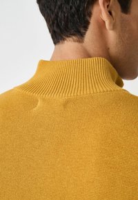Maglione a coste di colore senape con colletto alto. La texture è liscia con cuciture sottili al collo e sul retro, caratterizzato da un design aderente.