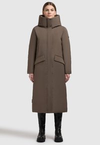 Lungo piumino marrone con collo alto, dotato di due tasche frontali e taglio dritto. Tessuto strutturato con dettagli di cuciture visibili.