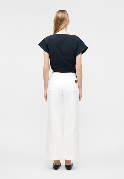 Γυναίκα με μακριά ξανθά μαλλιά που φορεί μαύρο τοπ με κοντά μανίκια, λευκό παντελόνι φαρδύ στα πόδια και μαύρα παπούτσια, στέκεται με την πλάτη προς τον θεατή.