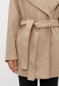 Beige belted kåpe med brede revers, iført over svarte bukser, viser venstre arm og hånd med lys beige neglelakk.