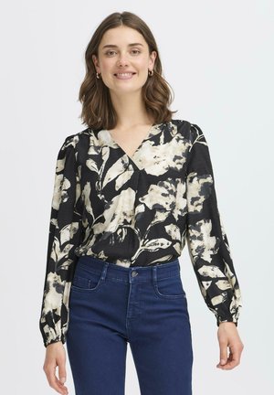 Femme aux cheveux bruns mi-longs sourit, portant un chemisier fleuri noir et crème et un jean bleu foncé, debout devant un fond uni.