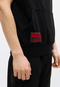 Μαύρο αμάνικο φούτερ με κουκούλα και μπροστινή τσέπη, με κόκκινο έμβλημα λογότυπου "Venum Tysons". Το υλικό φαίνεται απαλό· το τελείωμα είναι λίγο μακρύτερο στο πίσω μέρος.