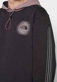 Sudadera negra con un cordón en gris claro y un forro de capucha púrpura en contraste. Presenta un logo cuadrado y acentos a rayas en la manga.