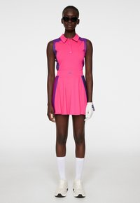 Rosa ärmlös polosklänning med krage och plisserad kjol, med lila sidodetaljer, i kombination med vita strumpor och beige sneakers.