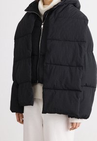 Personne portant plusieurs couches de doudounes noires sur un pull à col roulé crème et un pantalon blanc sur un fond uni.