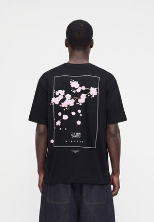 Άνδρας που φοράει μαύρο T-shirt με ροζ σχέδιο κερασιάς και ιαπωνικό κείμενο πλαισιωμένο στην πλάτη, σε συνδυασμό με σκούρο παντελόνι.