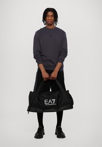 Borsa da palestra nera con logo bianco "EA7 Emporio Armani"; presenta manici doppi e un tessuto liscio e resistente. Modello che indossa un maglione grigio e pantaloni neri.