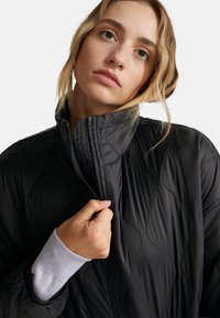 Černá prošívaná bunda s vysokým límcem a Zipovým uzávěrem, která vystihuje hladkou texturu a diamantový vzor. Ruka umístěná blízko zipu.