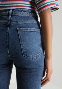 Jean en denim bleu taille haute avec une silhouette ajustée, présentant une poche arrière avec un détail de couture et une texture de tissu lisse.