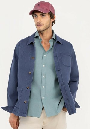 Hombre con gorra roja, chaqueta azul, camisa verde abotonada y pantalones beige, de pie con una mano en el bolsillo sobre un fondo liso.