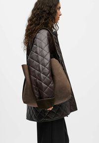 Sac hobo en daim marron avec une texture douce, une forme courbée et une grande ouverture. Assorti à une veste matelassée noire et un pantalon noir.